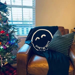 Navy DON'T BE SHITTY smiley blanket draped over a leather armchair beside a decorated Christmas tree