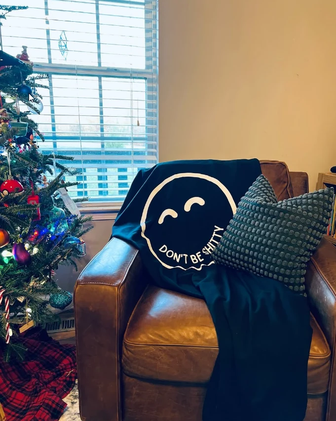 Navy DON'T BE SHITTY smiley blanket draped over a leather armchair beside a decorated Christmas tree