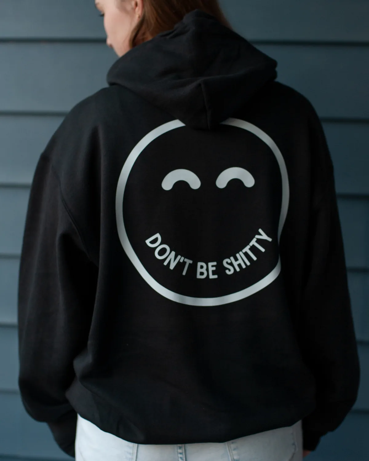 Back view of the black DON'T BE SHITTY smiley hoodie being worn, large smiley print across the shoulders