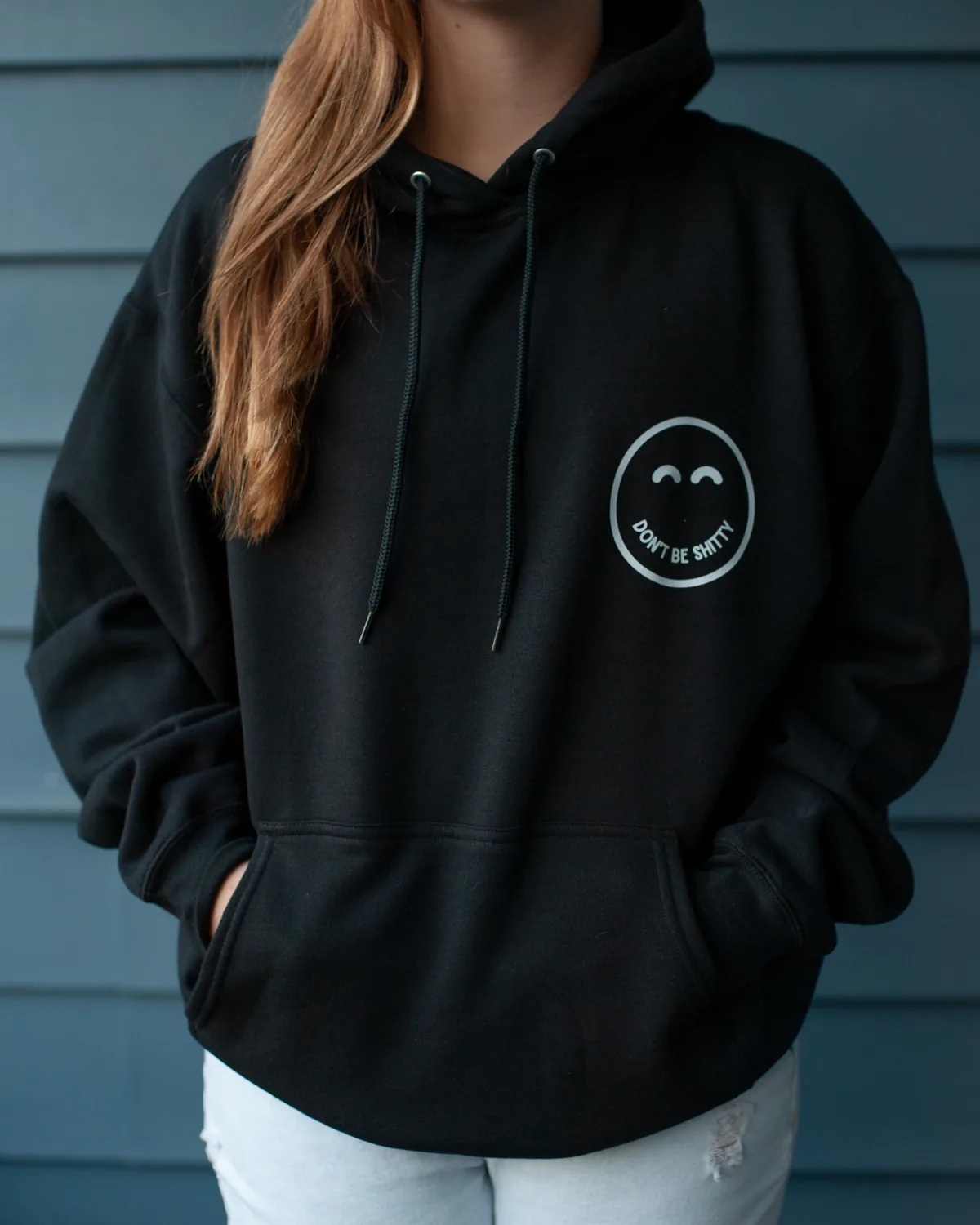 Woman wearing the black DON'T BE SHITTY smiley hoodie with her hands in the front pouch pocket