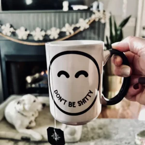 Hand holding the 15 oz DON'T BE SHITTY smiley mug with a black handle, fireplace and dog in the background