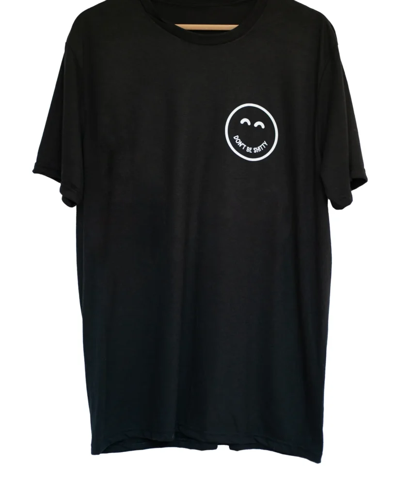 Black DON'T BE SHITTY smiley t-shirt on a hanger, small smiley logo on the left chest